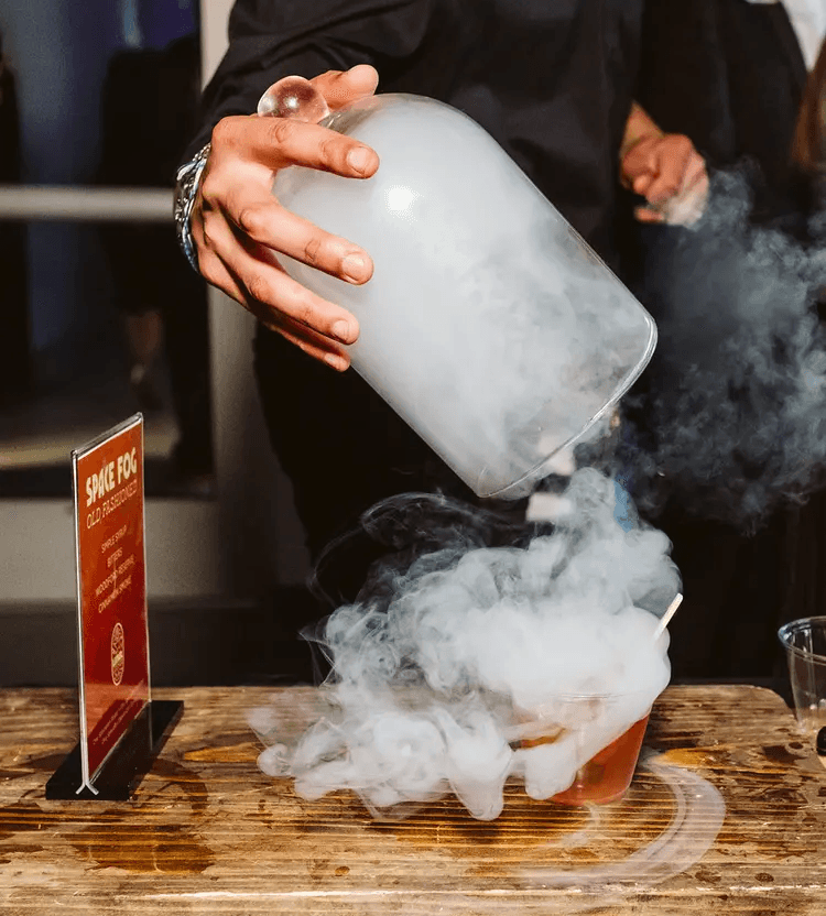 A person in a black shirt pours a smoky, fog-like vapor from a glass container over a drink on a wooden table, creating a dramatic “space fog” effect, while another person stands blurred in the background.